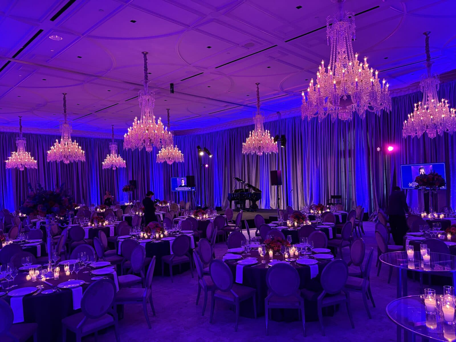 A ballroom under a canopy of crystal chandeliers with candlelit tables and a grand piano stage